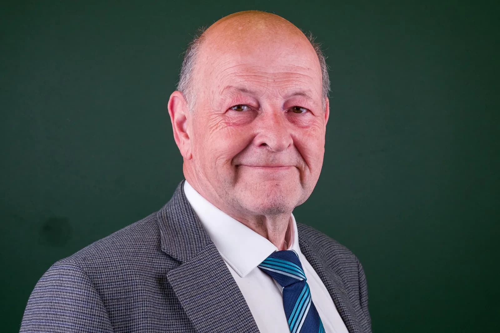 An older, balding man in a suit jacket, white shirt, and blue striped tie smiles slightly at the viewer.